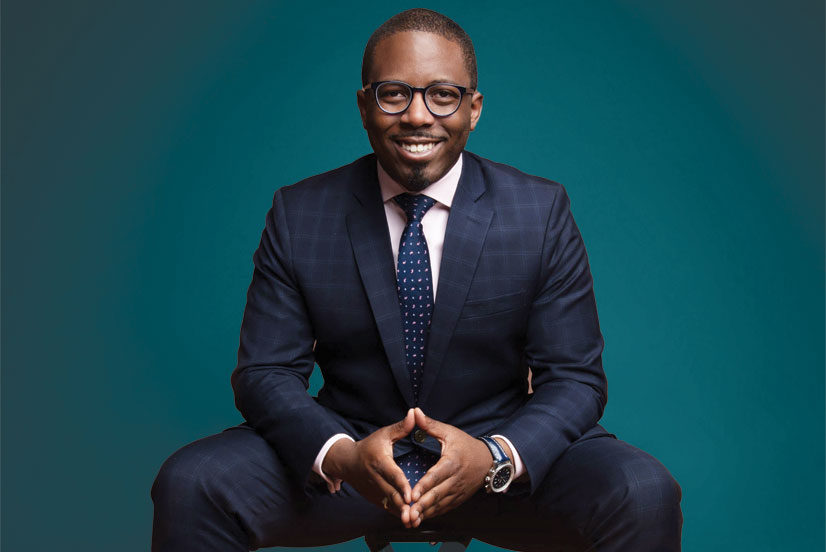 Aziz Garuba is a dark skinned man with a bright, wide smile grinning for the camera. He is sitting in a chair, leaned forward slightly with his hands clasped in front of him.