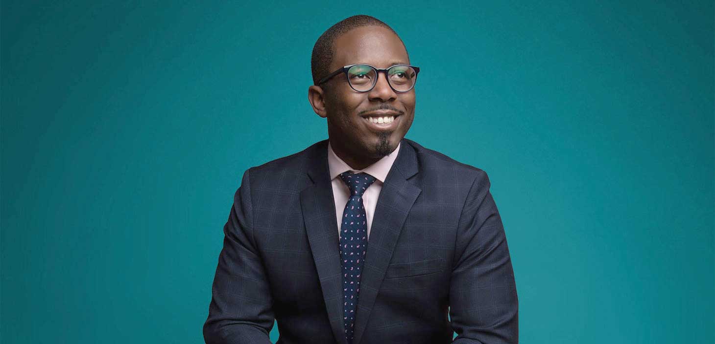 Aziz Garuba is a smiling dark skinned man sitting in front of a turquoise background.