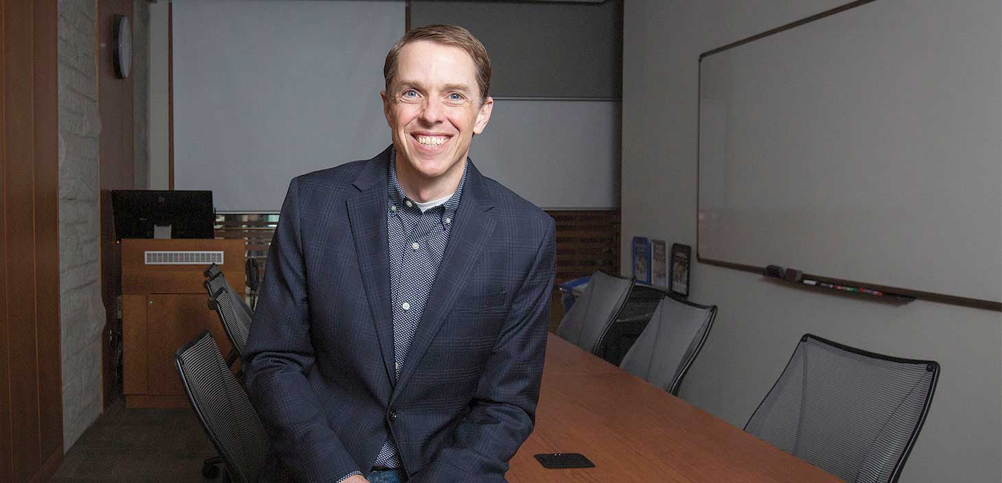 Jacob Brower stands beside the wall in a board room as he smiles wide for the camera. 